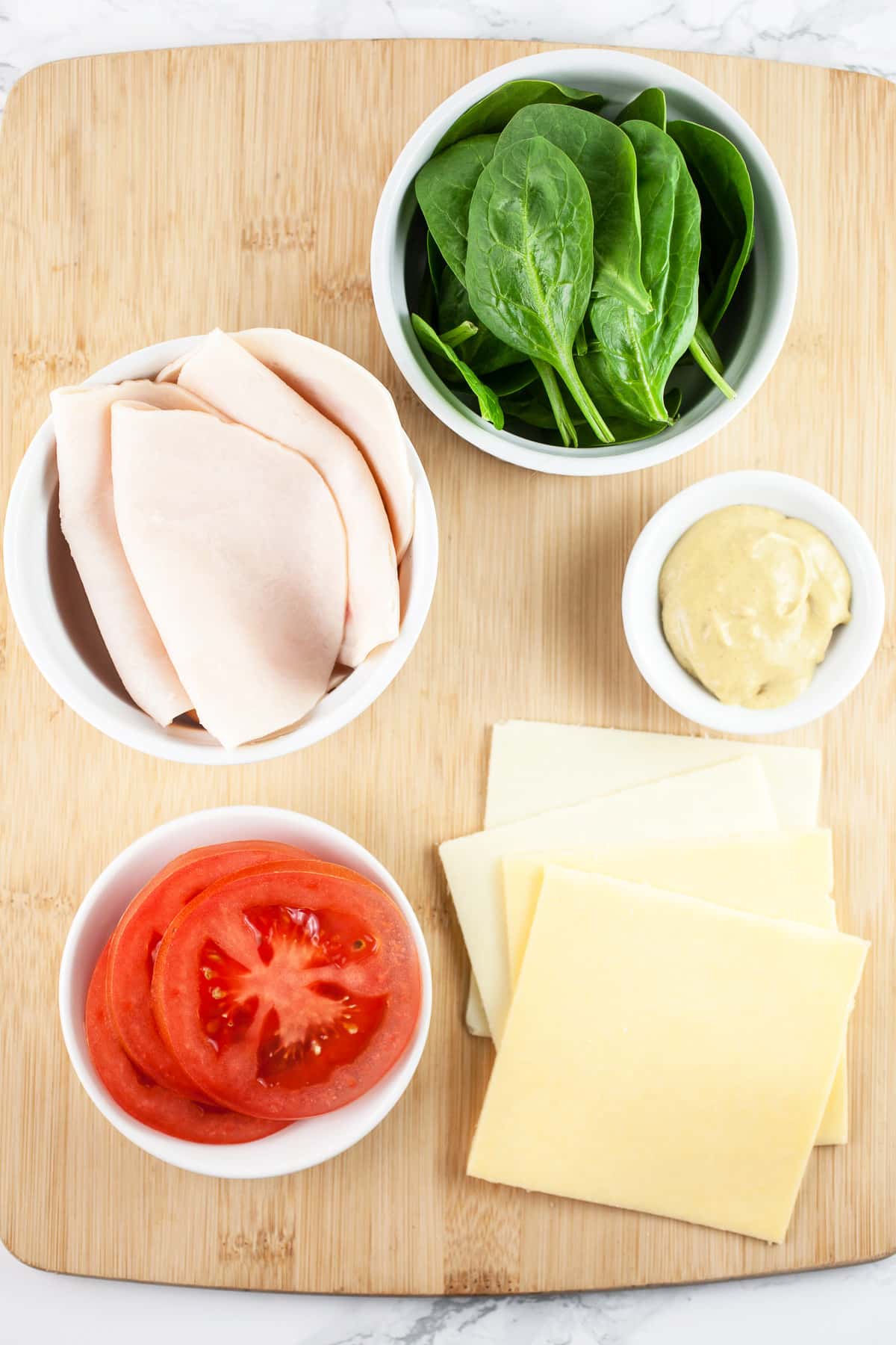 Sliced cheese, tomatoes, deli chicken, spinach, and mustard on wooden cutting board.