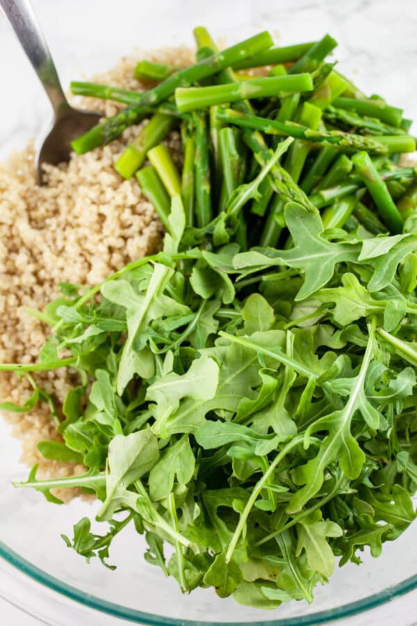 Cooked quinoa, blanched asparagus, and arugula in glass bowl with metal spoon.