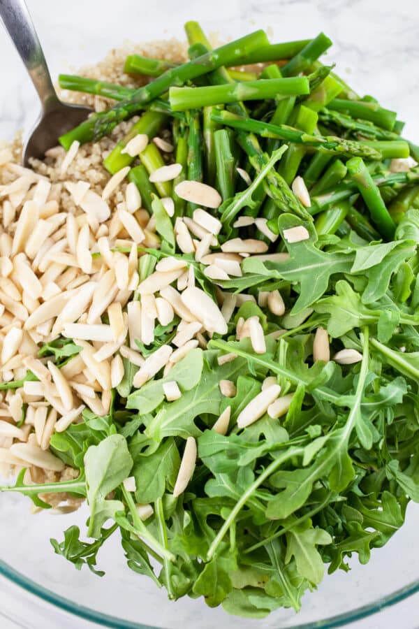 Cooked quinoa, blanched asparagus, arugula, and slivered almonds in glass bowl with metal spoon.