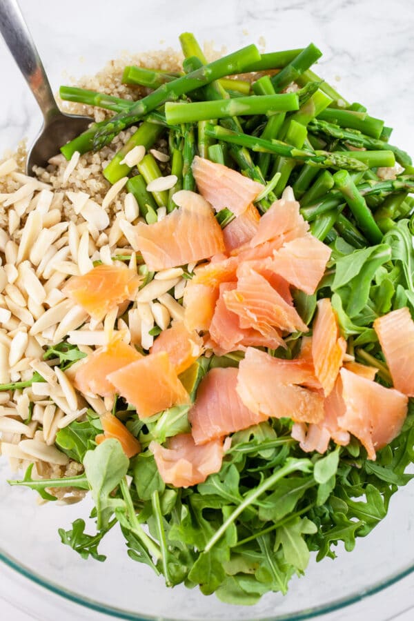 Cooked quinoa, blanched asparagus, arugula, slivered almonds, and smoked salmon in glass bowl with metal spoon.