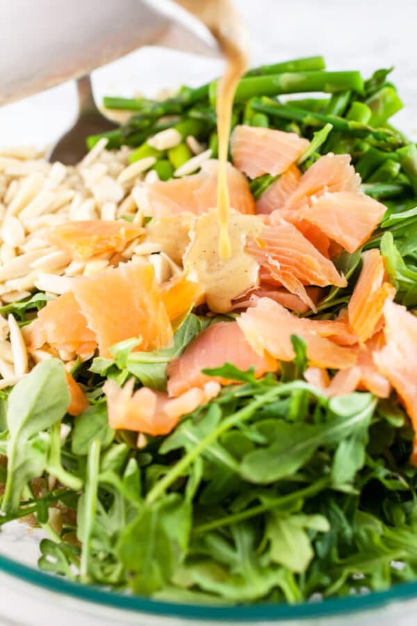 Parmesan vinaigrette poured onto smoked salmon arugula quinoa salad in large glass mixing bowl.