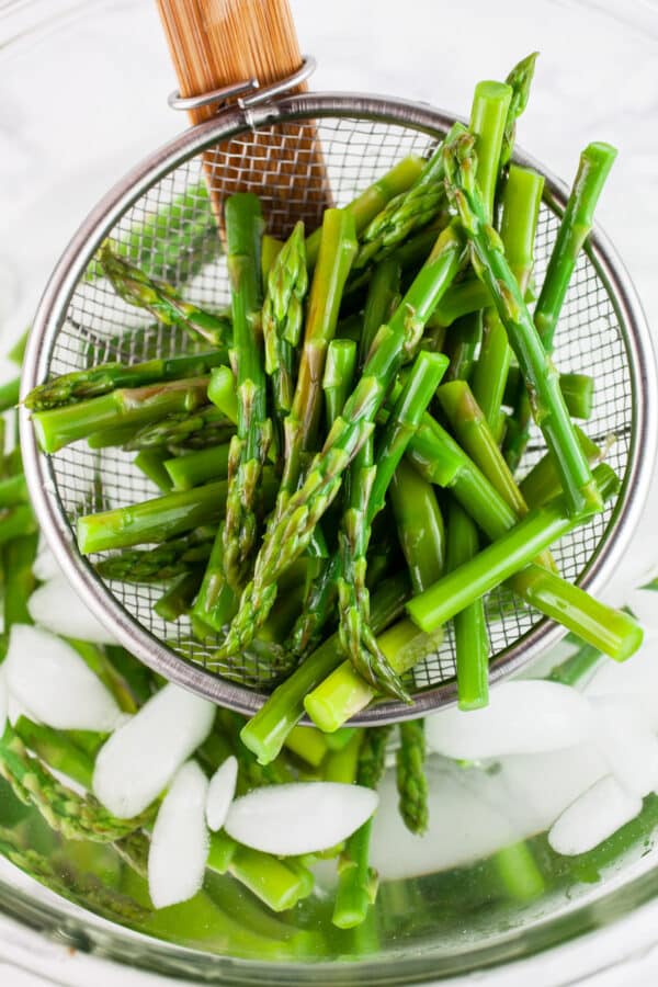 Blanched asparagus lifted from bowl of ice water on metal strainer.