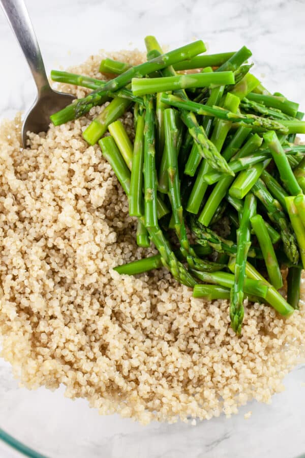 Cooked quinoa and blanched asparagus in glass bowl with metal spoon.