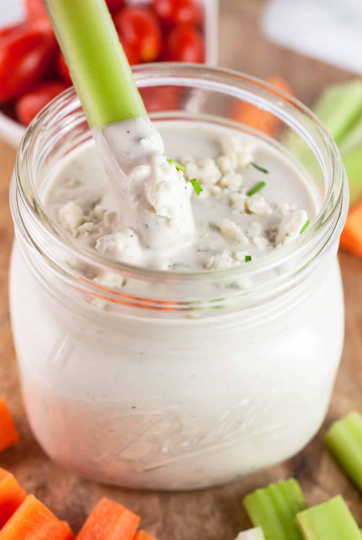 Celery stick dipped into jar of blue cheese dressing sitting on a wooden board with raw veggies.