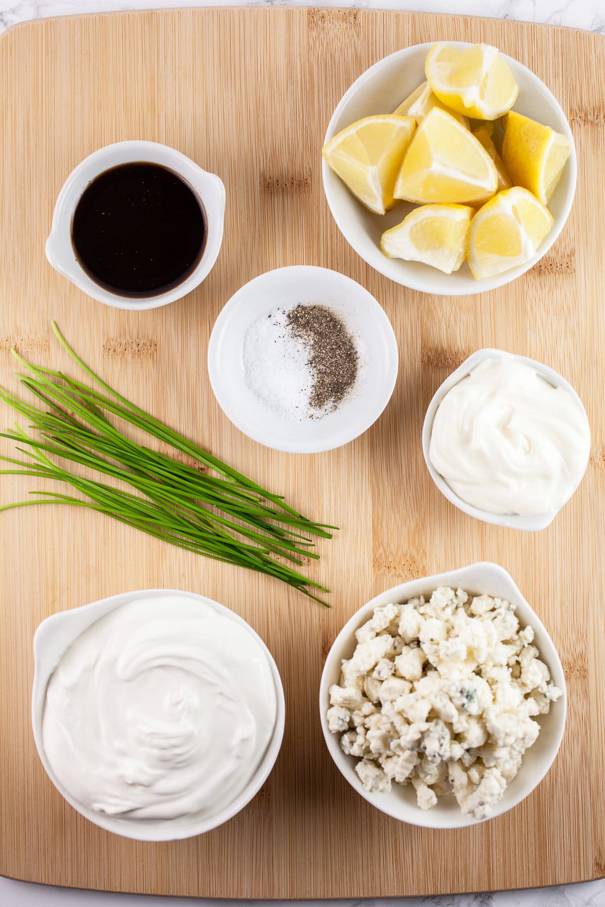 Blue cheese crumbles, sour cream, mayo, chives, Worcestershire sauce, lemon wedges, and salt and pepper on wooden cutting board.