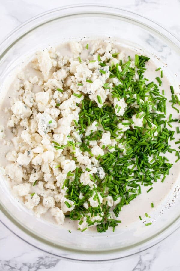 Blue cheese crumbles and minced chives added to creamy dressing in glass bowl.