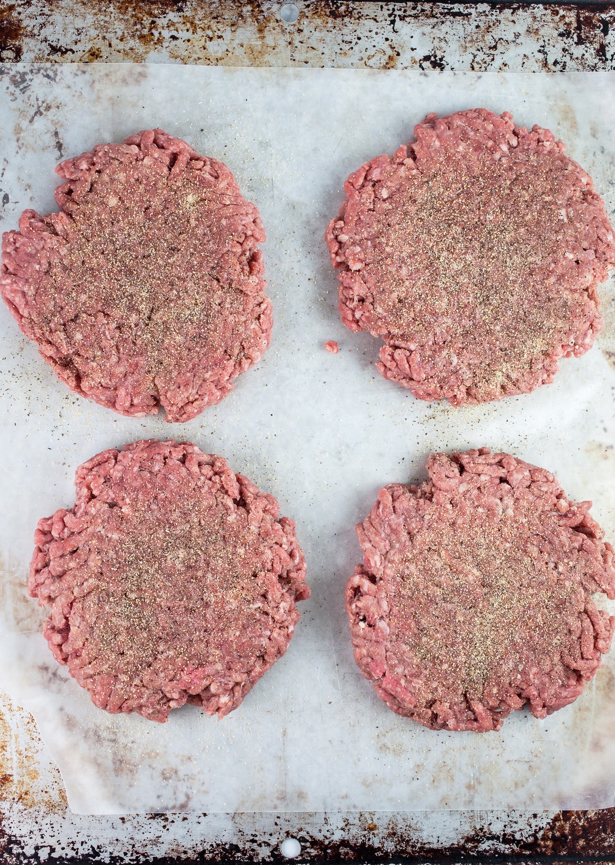 Seasoned raw ground beef patties on metal pan.