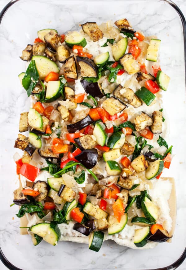 Sautéed vegetables over lasagna noodles and ricotta cheese mixture in glass baking dish.