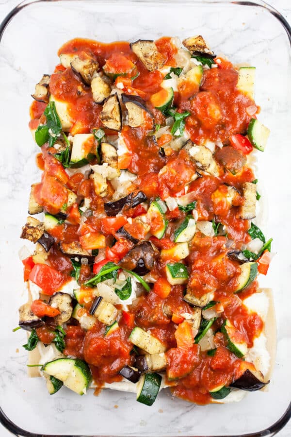 Marinara sauce over sautéed vegetables, lasagna noodles, and ricotta cheese mixture in glass baking dish.