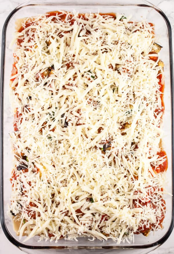 Uncooked vegetable lasagna with shredded cheese in glass baking dish.