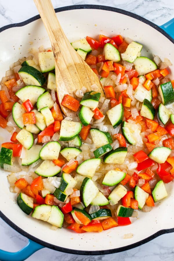 Garlic, onions, zucchini, and red bell peppers sautéed in skillet with wooden spoon.
