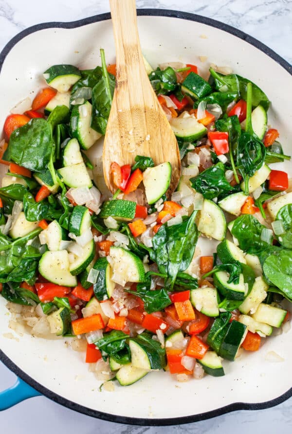 Garlic, onions, zucchini, red bell peppers, and spinach sautéed in skillet with wooden spoon.