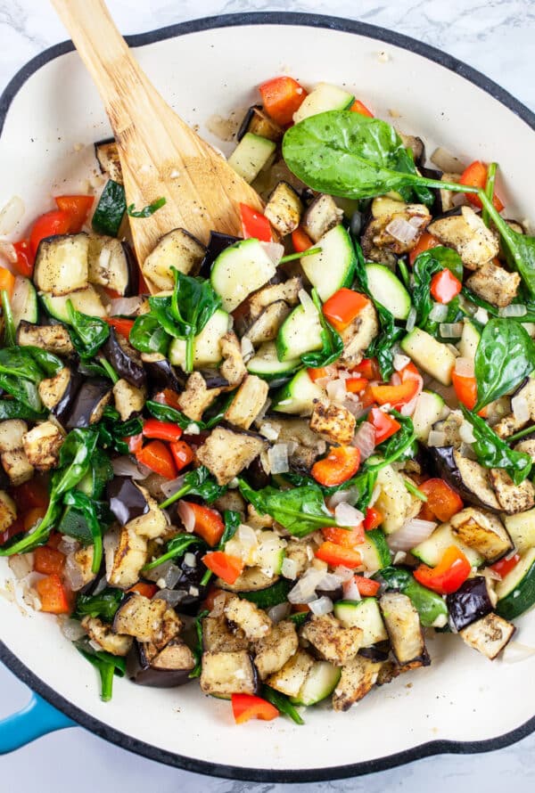 Garlic, onions, zucchini, red bell peppers, spinach, and eggplant sautéed in skillet with wooden spoon.