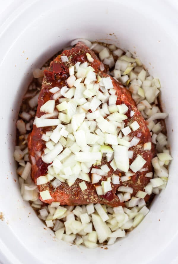 Uncooked beef chuck roast with diced onions in slow cooker.