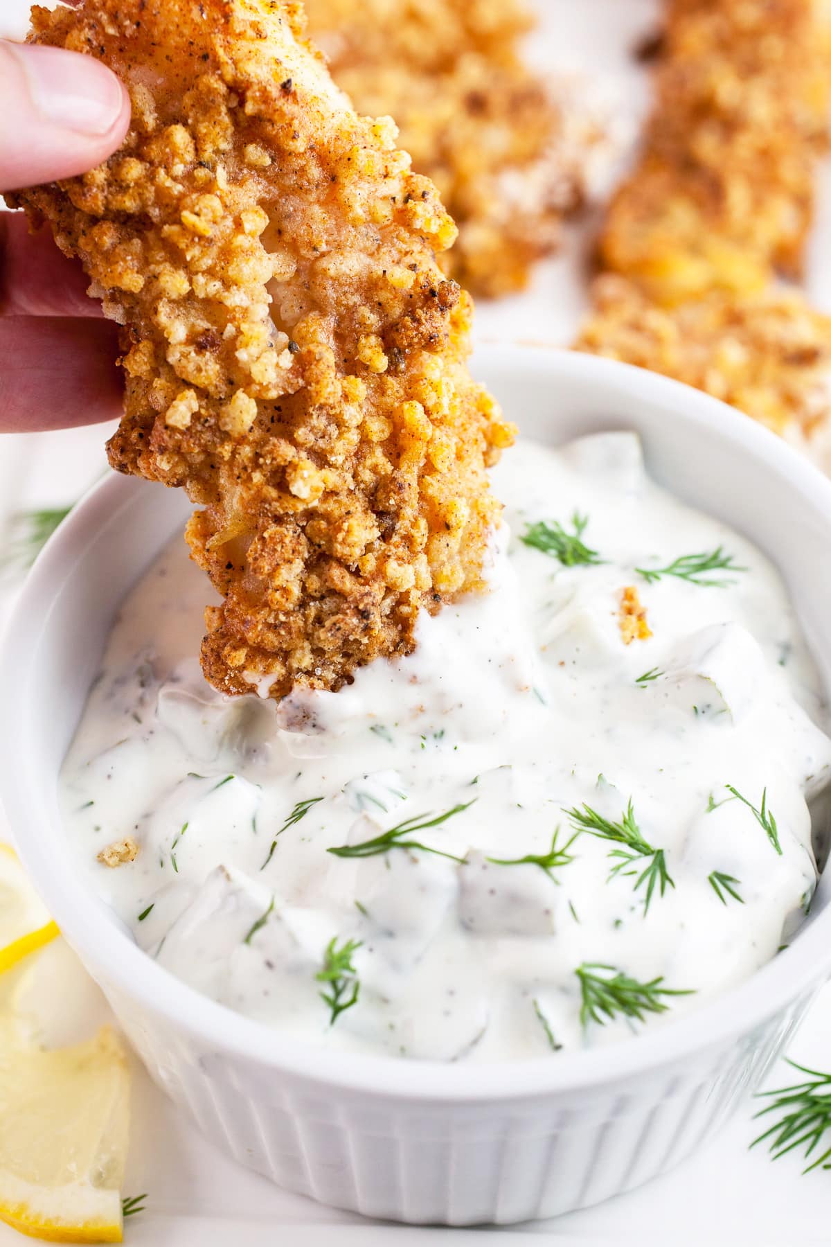 Fish finger dipped into small white bowl of dill tartar sauce.