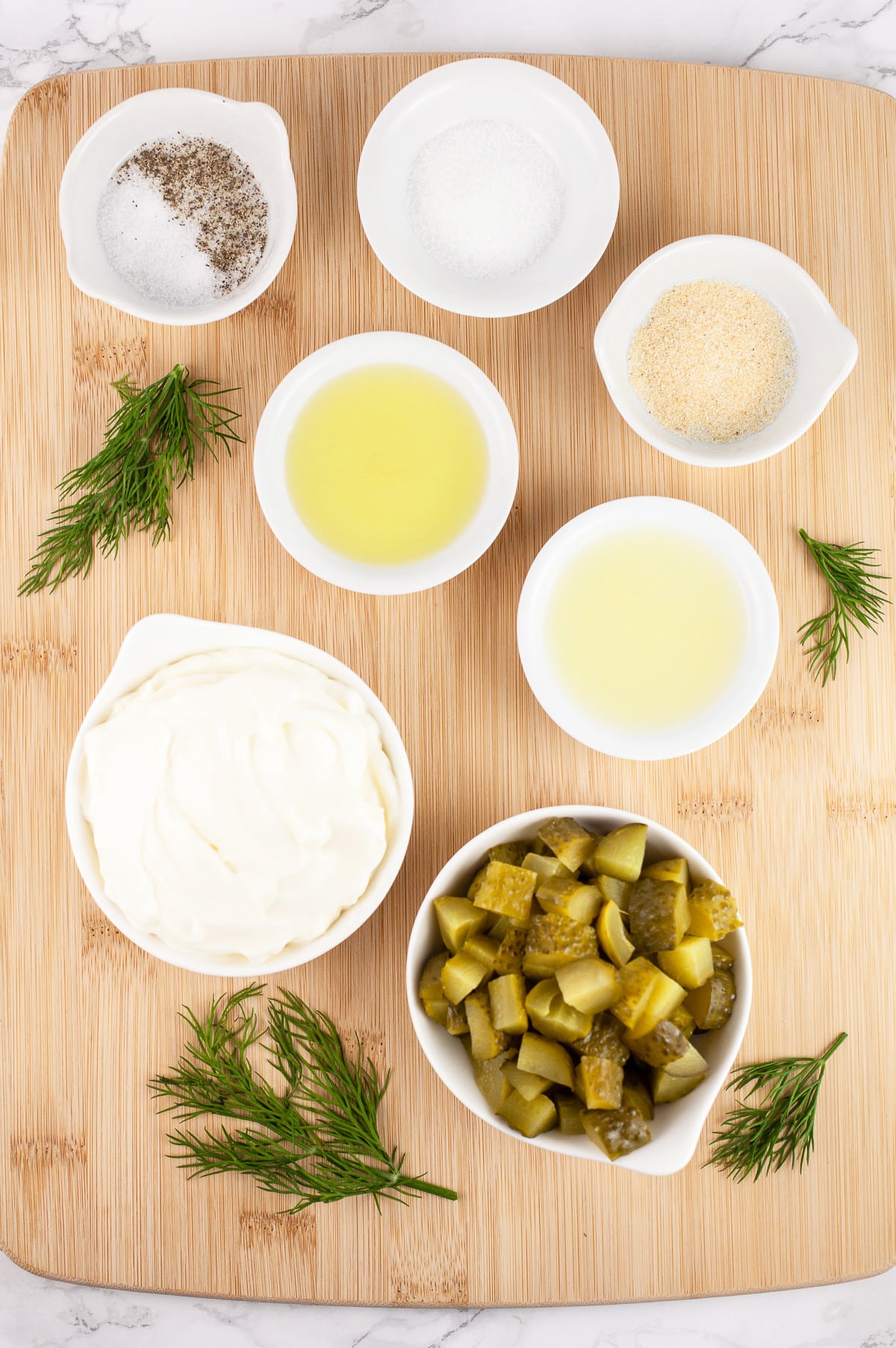Chopped dill pickles, mayo, lemon juice, pickle juice, garlic powder, sugar, salt and pepper, and fresh dill on wooden cutting board.