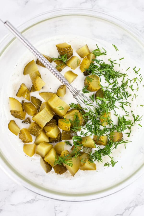 Diced dill pickles and fresh dill added to mayo in glass bowl with whisk.
