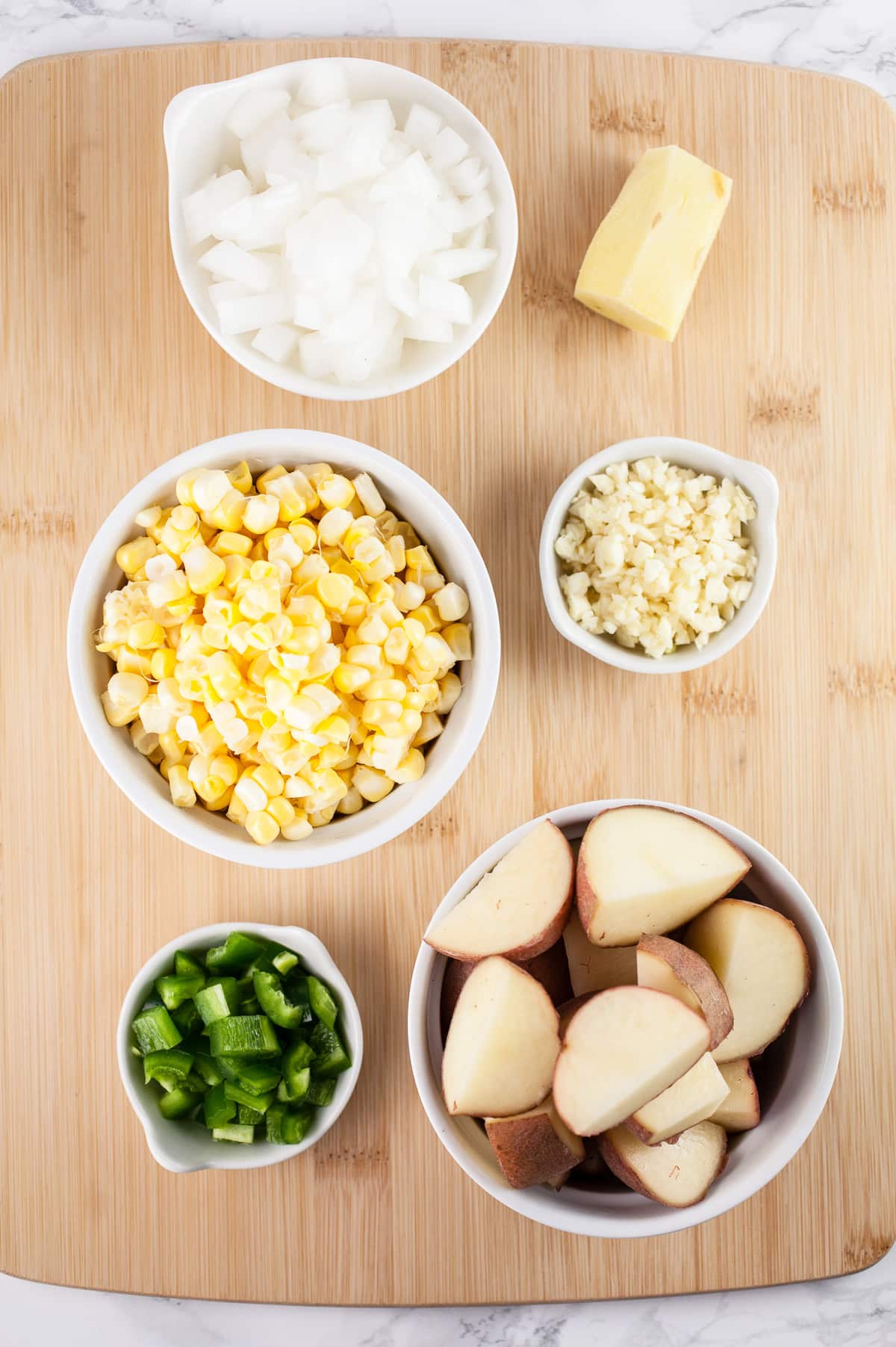 Minced jalapenos, diced red potatoes, sweet corn, garlic, onions, and ginger root on wooden cutting board.