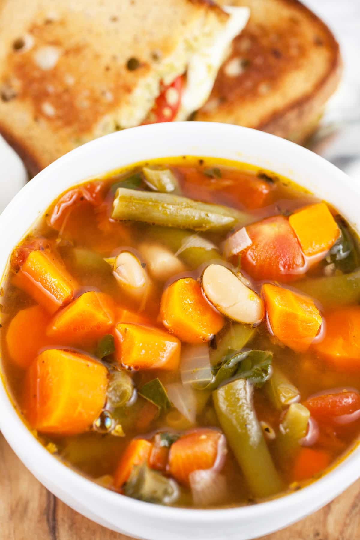 This Vegetable Soup and Pesto Grilled Cheese makes a great lunch or dinner any time of year! This hearty homemade Italian vegetable soup is easy to make and pairs perfectly with a pesto grilled cheese sandwich. This sandwich is full of melted mozzarella cheese, tomato, and basil pesto. A soup and sandwich is one of the best pairings ever! It makes the perfect simple meal. #soup #sandwich #vegetablesoup #pesto #grilledcheese #vegetarian #lunch #dinner Homemade vegetable soup in white bowl in front of pesto grilled cheese sandwich cut in half.