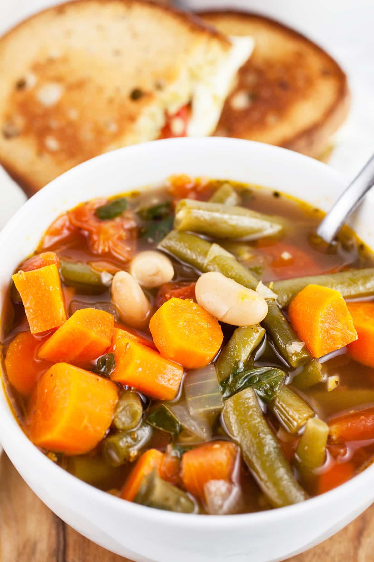 This Vegetable Soup and Pesto Grilled Cheese makes a great lunch or dinner any time of year! This hearty homemade Italian vegetable soup is easy to make and pairs perfectly with a pesto grilled cheese sandwich. This sandwich is full of melted mozzarella cheese, tomato, and basil pesto. A soup and sandwich is one of the best pairings ever! It makes the perfect simple meal. #soup #sandwich #vegetablesoup #pesto #grilledcheese #vegetarian #lunch #dinner Vegetable soup in white bowl with spoon in front of pesto grilled cheese cut into halves.