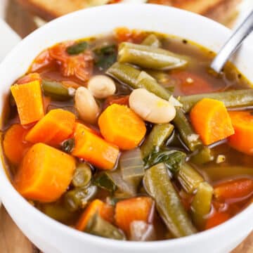 Vegetable soup in white bowl with spoon in front of pesto grilled cheese cut in half.