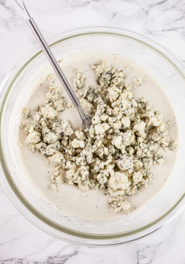 Blue cheese crumbles added to creamy dressing in glass bowl with small whisk.