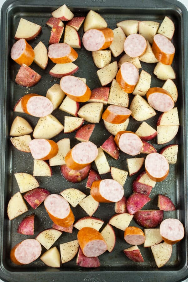 Uncooked potatoes and sliced sausages on baking sheet.