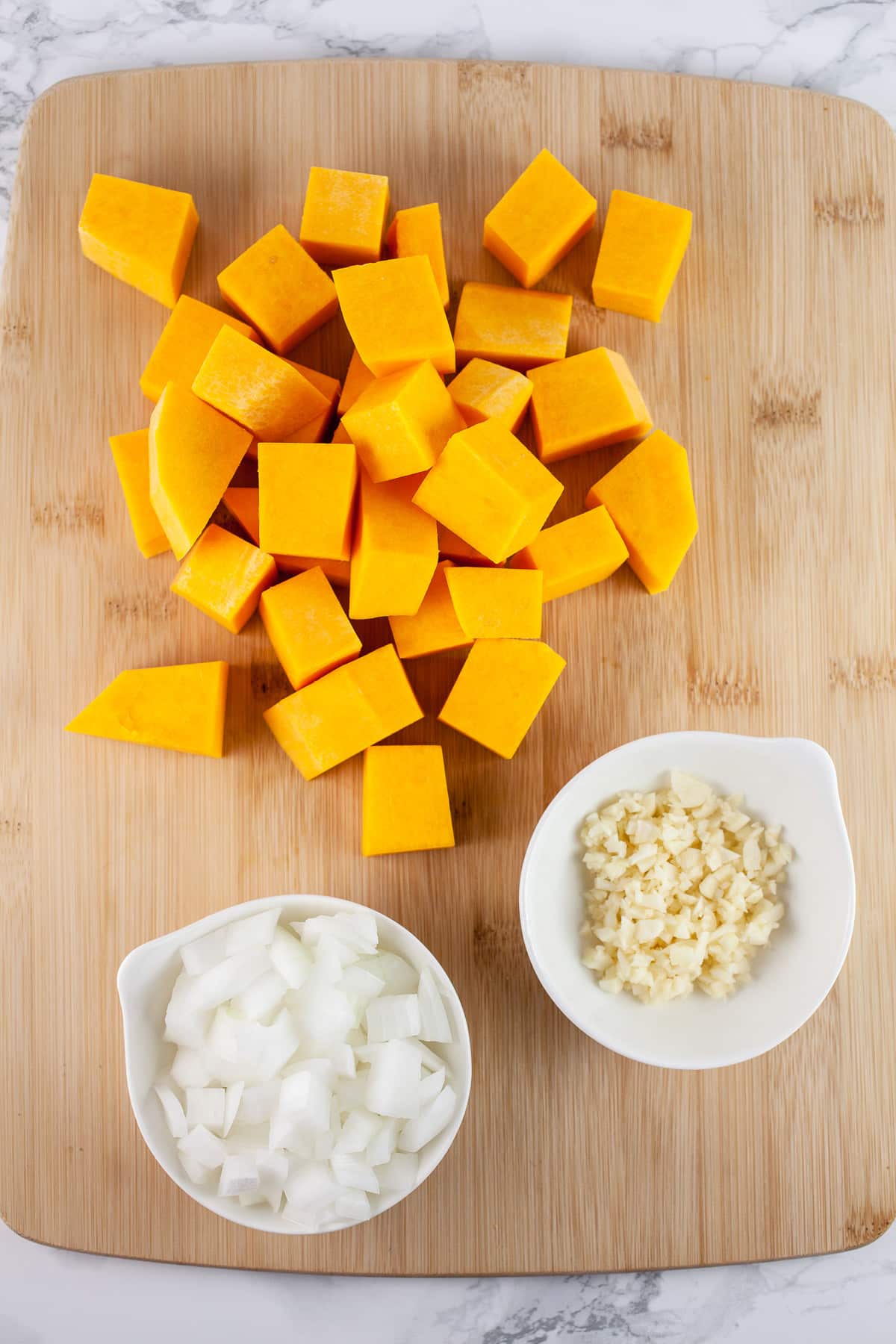 Minced garlic and onions and butternut squash cubes on wooden cutting board.
