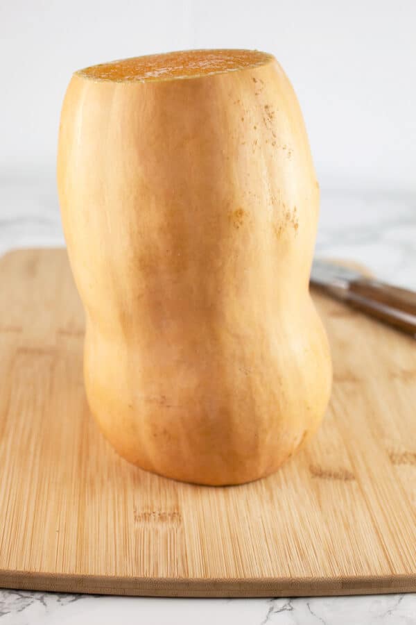Butternut squash with top and bottom removed on wooden cutting board with knife.