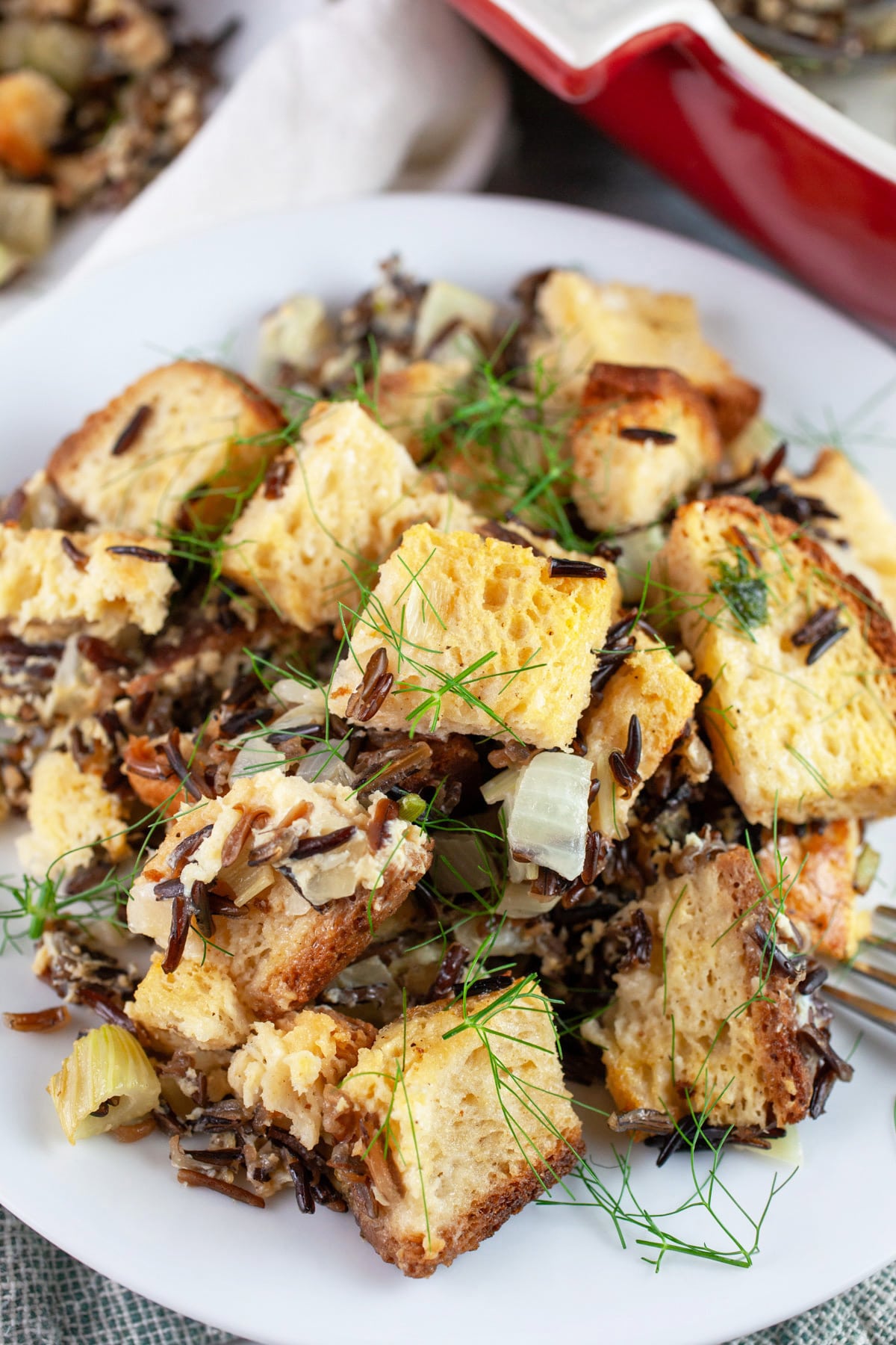 This Wild Rice and Fennel Stuffing recipe makes the perfect Thanksgiving side dish! It's vegetarian and one of the best healthy comfort foods for the holidays. This moist homemade stuffing is made with bread cubes, wild rice, and fennel and then baked in the oven. Everyone will love this unique take on a traditional classic. Stuffing made from scratch is the best! #stuffing #rice #fennel #vegetarian #thanksgiving #sides #recipes Wild rice fennel stuffing on white plate next to red casserole dish.