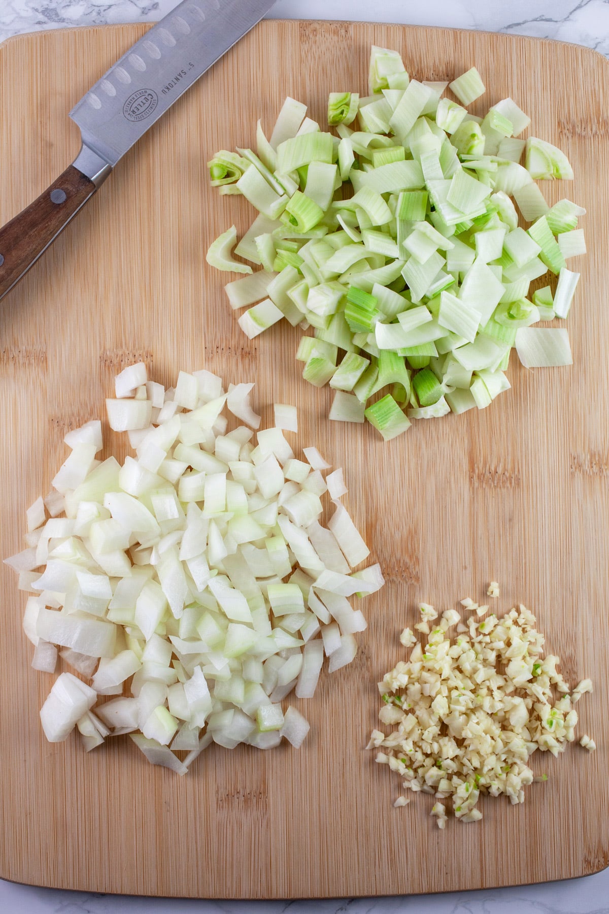 Minced garlic, onions, and fennel on wooden cutting board with knife.
