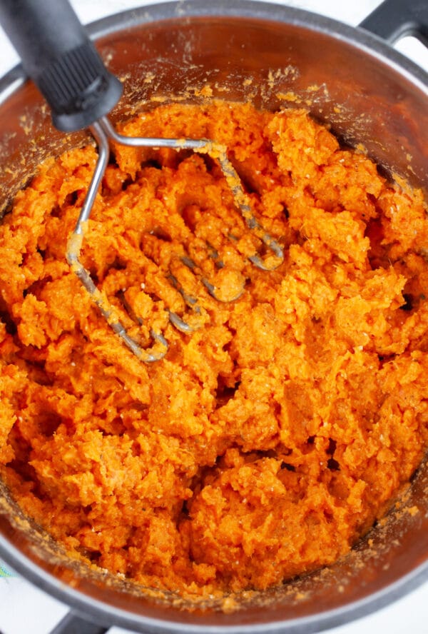 Mashed sweet potatoes in pot with potato masher.
