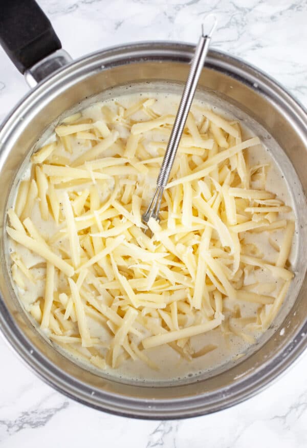 Grated Gruyere cheese added to creamy mixture in sauce pan with small whisk.