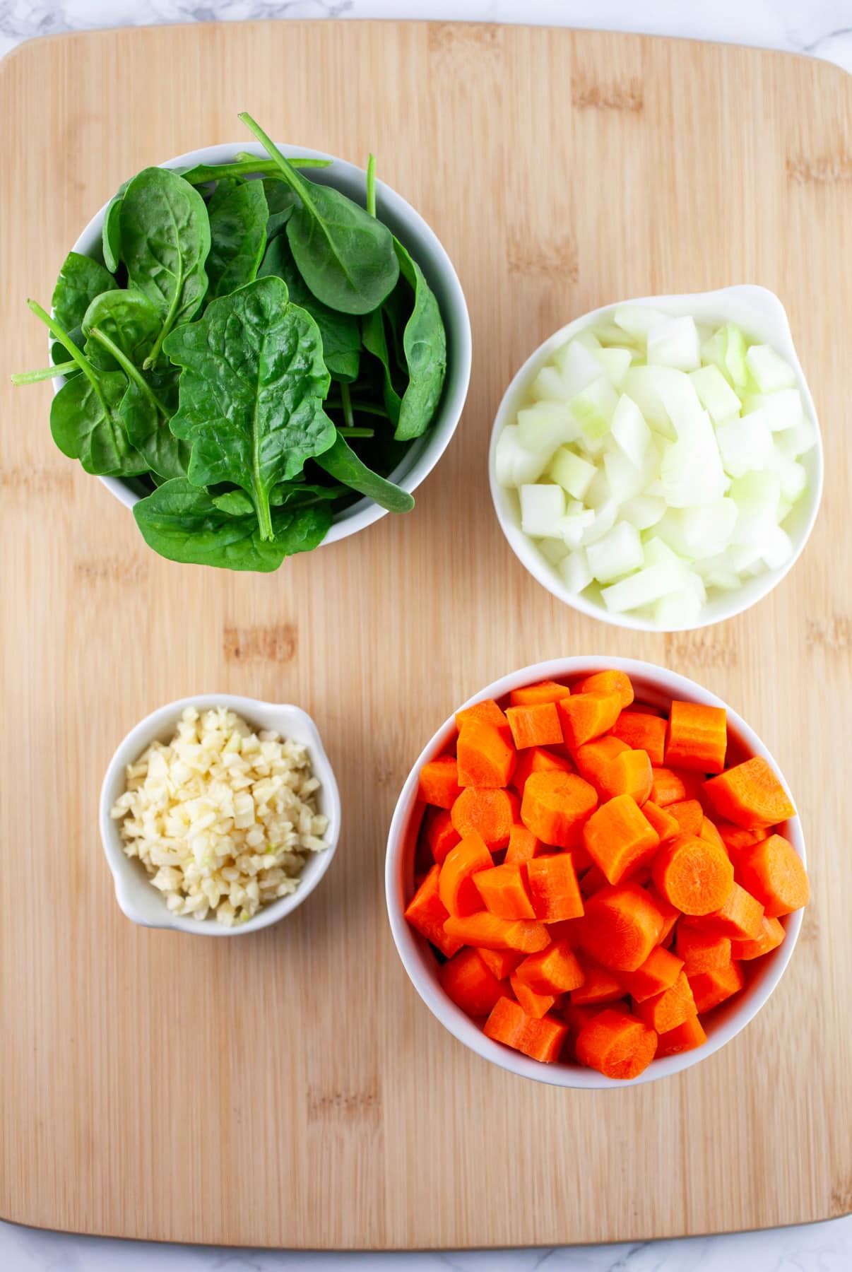 Minced garlic, onions, chopped carrots, and fresh spinach in white bowls on wooden cutting board.