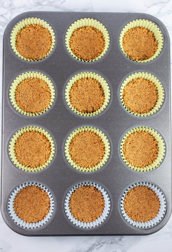 Unbaked graham cracker crusts in muffin tin lined with cupcake liners.