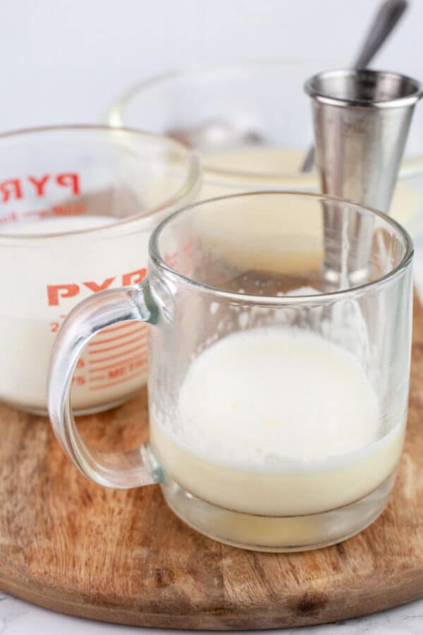 Tom and Jerry batter, warmed milk, and rum added to glass coffee mug and bowl of batter on wooden serving tray.