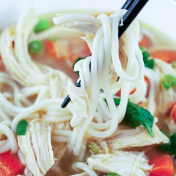 Chopsticks lifting Asian chicken noodle soup from white bowl.