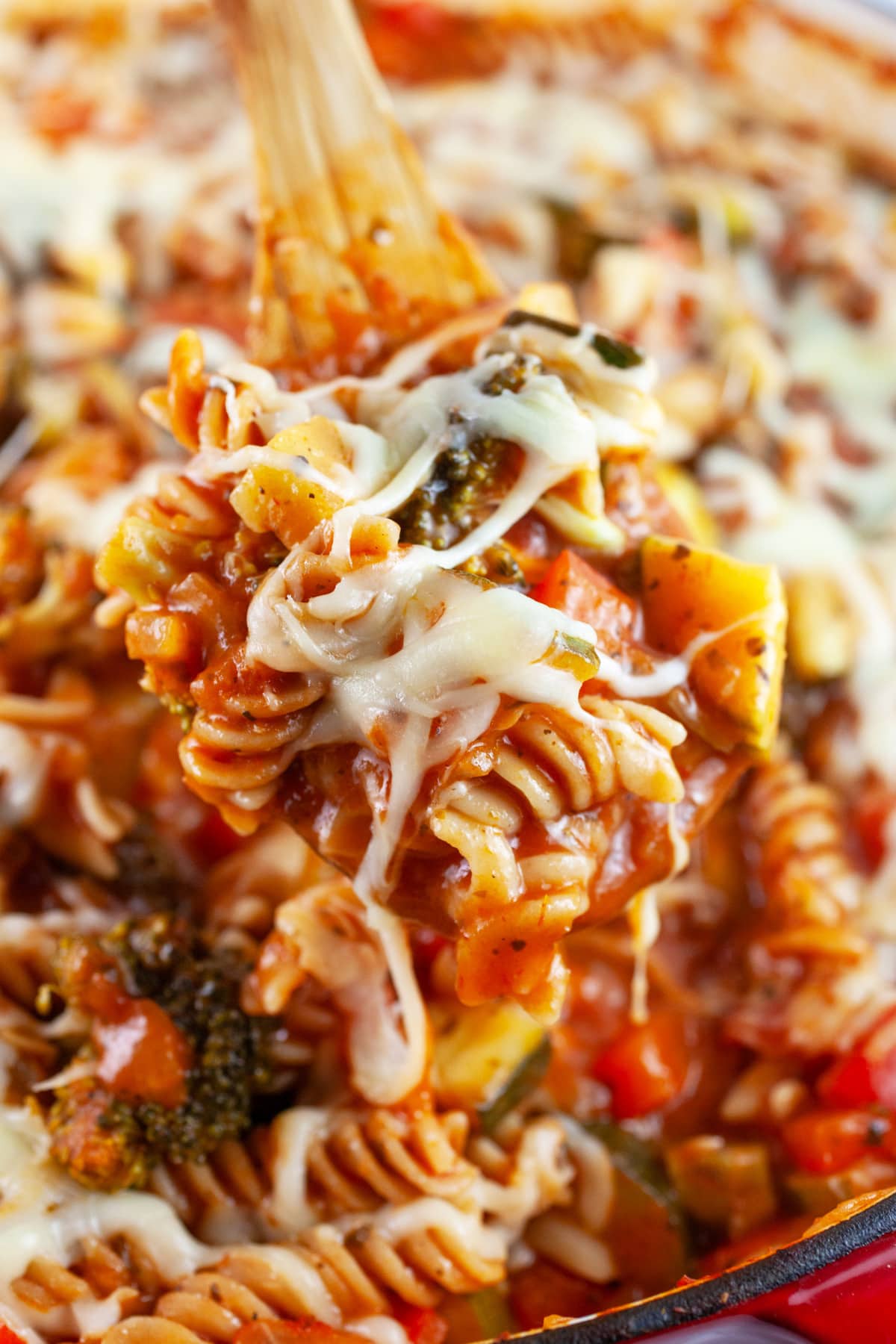 Scoop of cheesy veggie pasta lifted from skillet with wooden spoon.