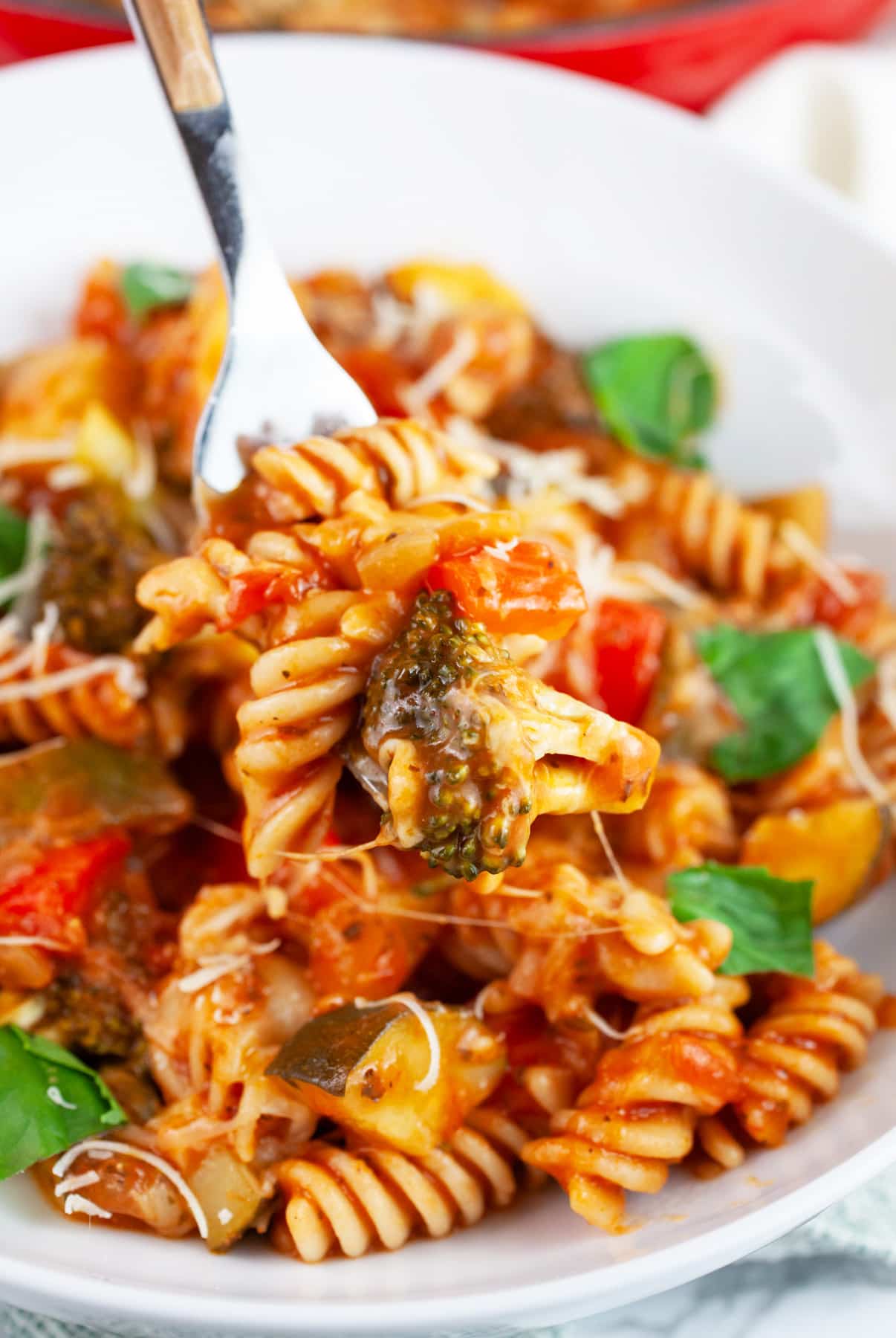 Forkful of vegetable pasta garnished with fresh basil lifted from white bowl.