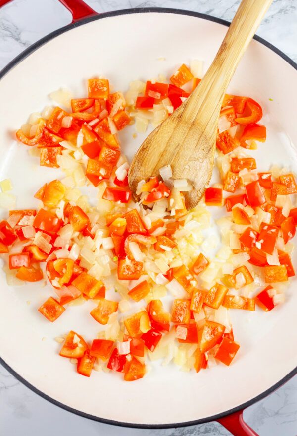 Minced garlic, onions, and red bell peppers sauteed in skillet with wooden spoon.
