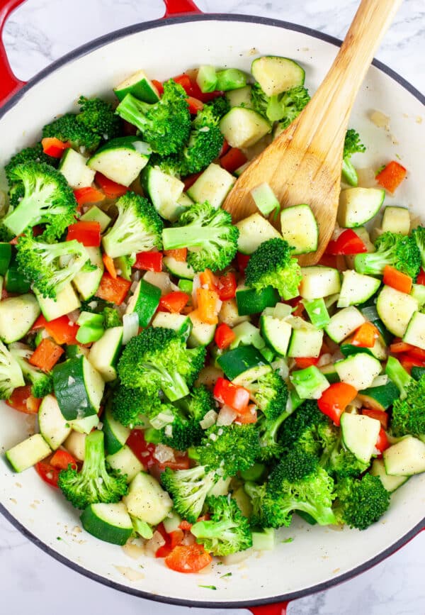 Garlic, onions, red bell peppers, zucchini, and broccoli florets sauteed in skillet with wooden spoon.