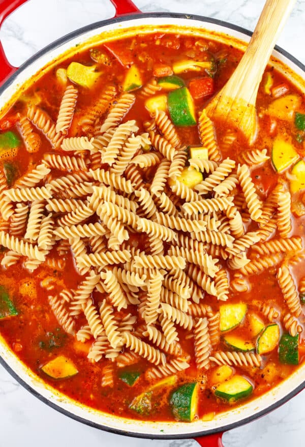 Sauteed vegetables and uncooked pasta with marinara sauce in skillet with wooden spoon.