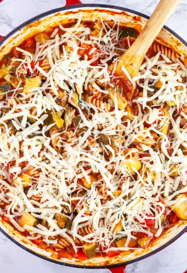 Veggie pasta with unmelted shredded mozzarella cheese in skillet with wooden spoon.