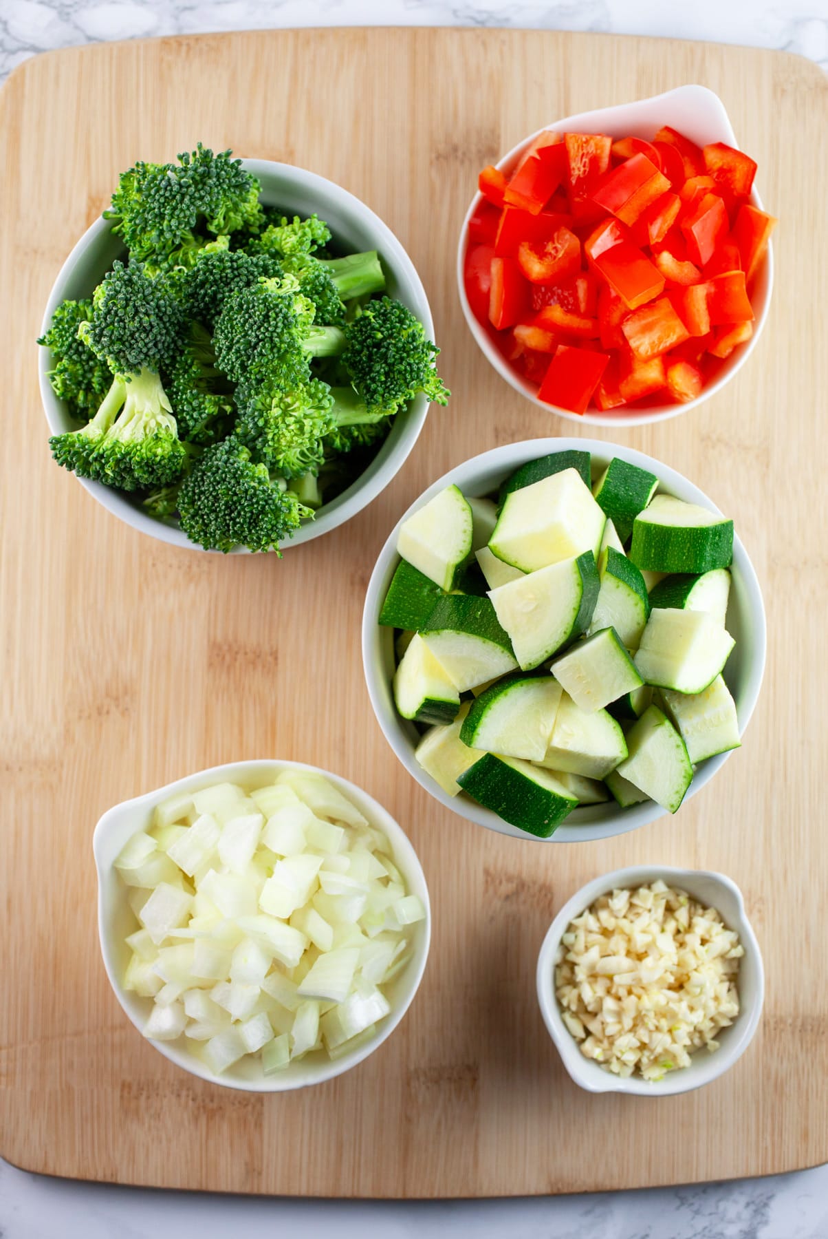 Minced garlic and onions, diced red bell peppers and zucchini, and broccoli florets in small white bowls on wooden cutting board.
