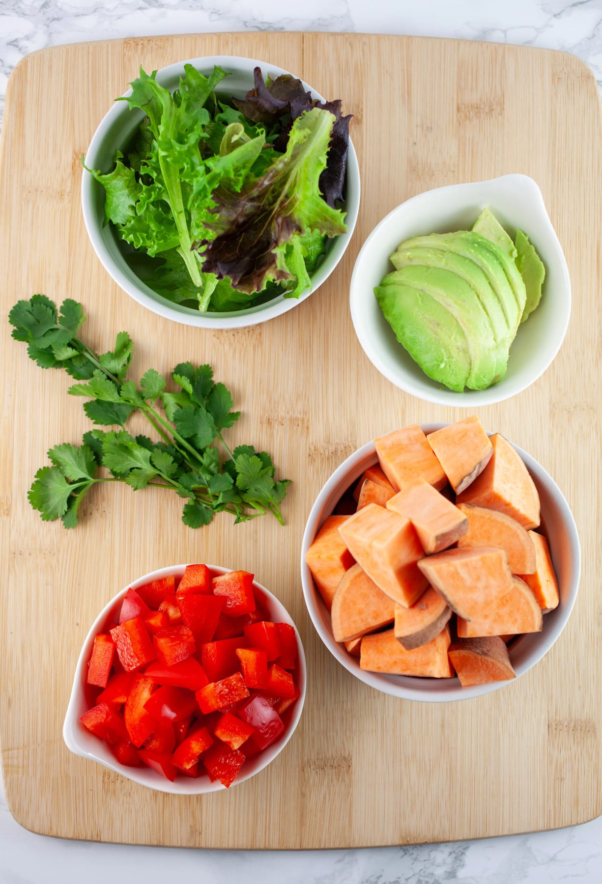 Diced sweet potatoes, red bell peppers, sliced avocado, greens, and cilantro on wooden cutting board.