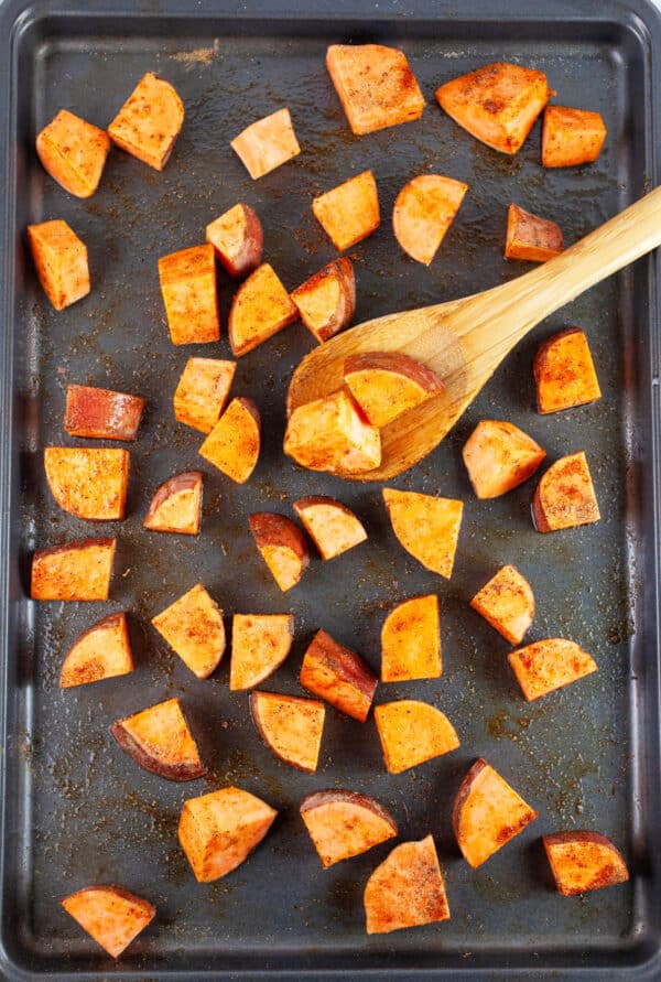 Uncooked chopped sweet potatoes in olive oil and spices on baking sheet with wooden spoon.