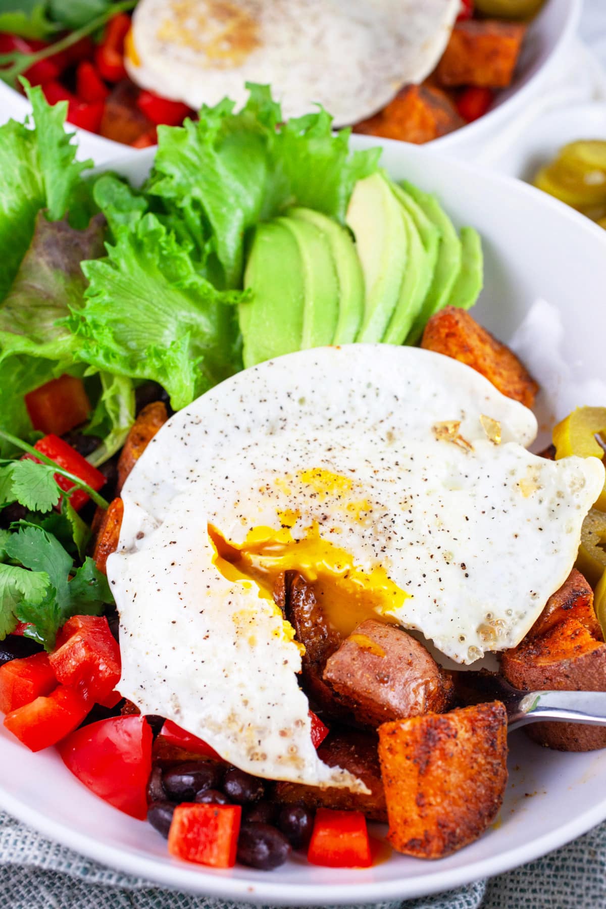 These Healthy Sweet Potato Breakfast Bowls are fast, easy to make, and full of protein. They're great for breakfast, lunch, or dinner. These breakfast bowls are full of roasted sweet potatoes, black beans, and topped with an egg. I love the savory Southwest flavors in this quick and easy meal. Here's to fresh veggies and clean eating! #sweetpotatoes #blackbeans #breakfastbowls #vegetarian Sweet potato breakfast bowls with egg cut in half by fork in white bowl.