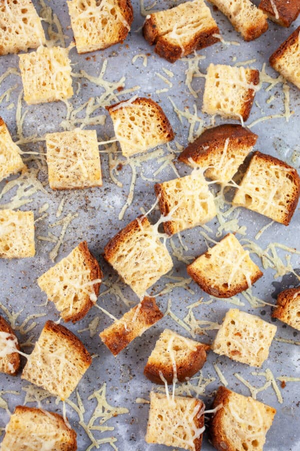 Toasted bread cubes with grated Asiago cheese on baking sheet lined with parchment paper.
