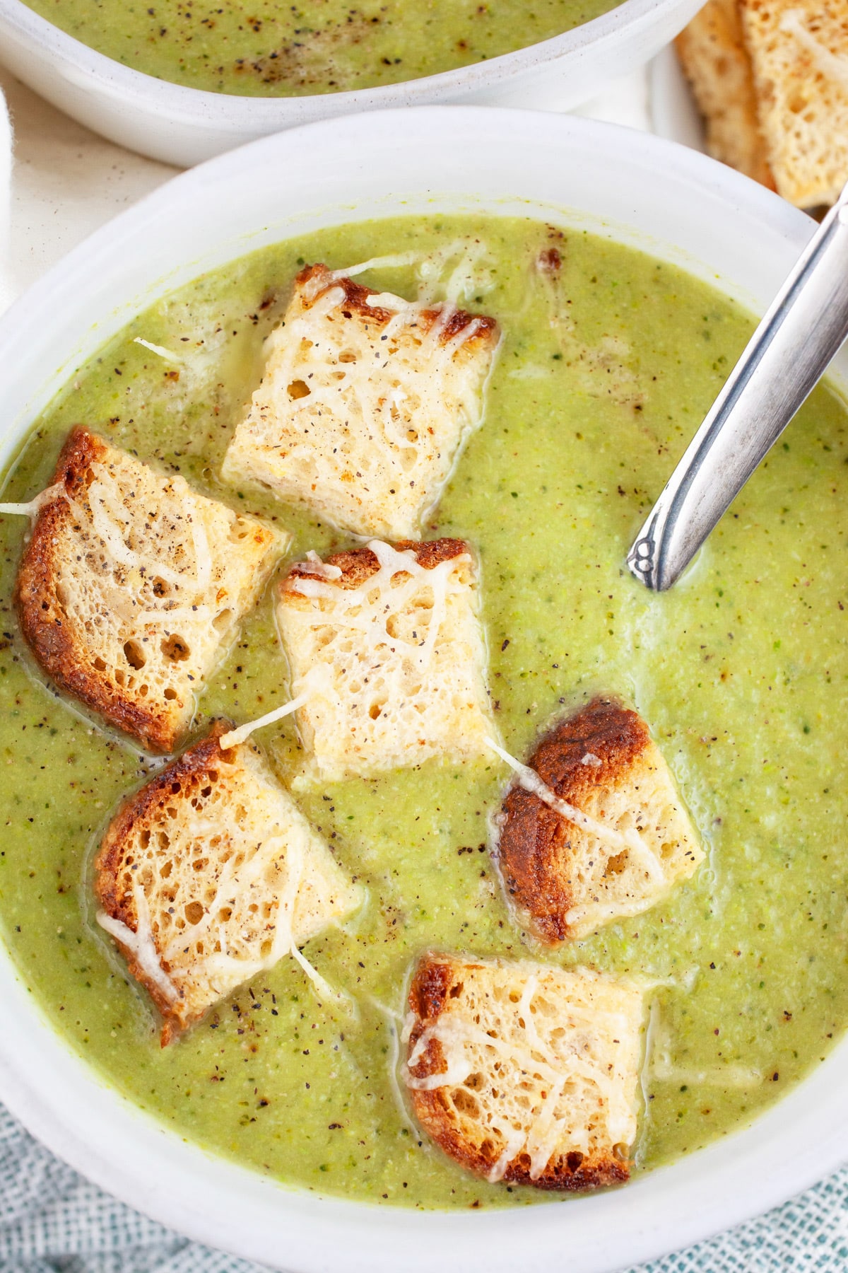 Broccoli asparagus soup with toasted Parmesan bread cubes in white bowl with spoon.