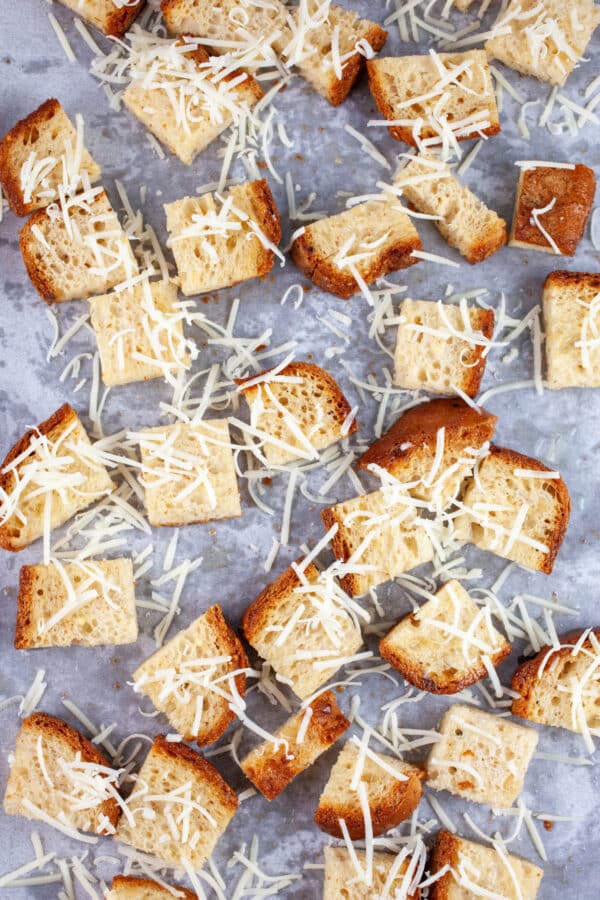 Bread cubes tossed in olive oil with grated Asiago cheese on baking sheet lined with parchment paper.